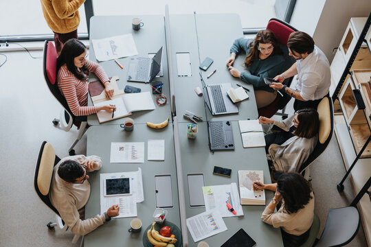 Coworkers collaborate and work together in a modern office. They discuss tasks, use laptops, and smile. This successful team represents a multicultural and multi-generational workforce. - Powered by Adobe