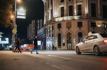 Two pedestrians casually crossing a well-lit city street at night with cars passing by and...
