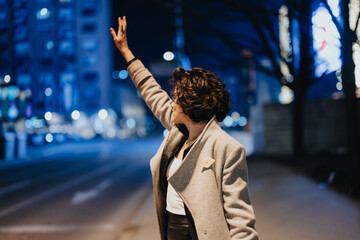 An adult woman signals for a cab on a bustling city street at night, with ambient light effect...
