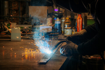 Master is doing welding at his workplace in the workshop, while sparks are flying all around, they are wearing a protective helmet.