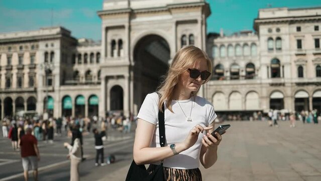 Picture of pretty young woman staying on the street holding phone in hands. Stylish blonde looking away is exploring new city in Milan Cathedral Square, Italy