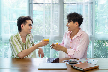 Cute Asian LGBT gay man couple, spend time together, with tablet, and laptop, on table. Holding orange juice glass.  Healthy lifestyle and work from home.