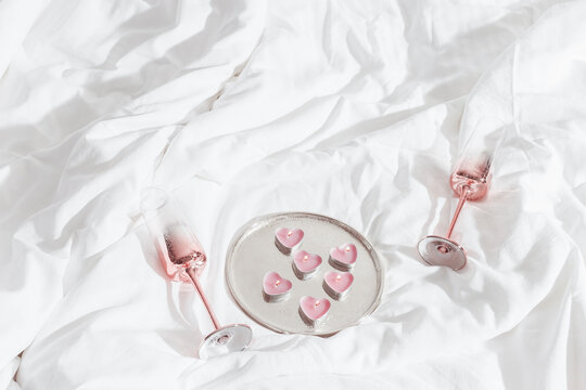 Pink shining champagne glasses and burning candles as hearts on bed cloth. Lifestyle aesthetic photo, star filter. Valentine's Day, love concept, romance meeting. Sparkling wine in wineglasses.