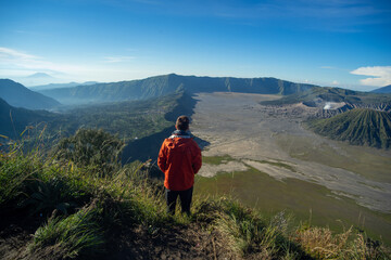 A person in an orange jacket admiring the view of a vast crater from a mountain summit