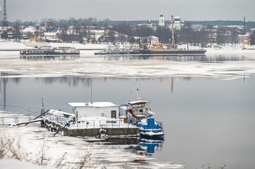 A small ship and boats on a small pier, covered with snow, on a river covered with ice and snow.