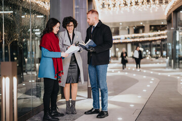Three young people stand together, reviewing documents amidst an illuminated evening shopping area...