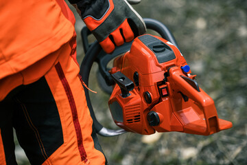 Close up view of an hand holding a chainsaw. Lumberjack at work wears orange personal protective...