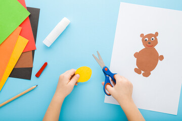 Little child hands holding scissors, cutting application paper and creating brown teddy bear on white sheet. Light blue table background. Pastel color. Point of view shot. Closeup. Top down view.