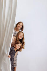 Three girls peek out from behind a curtain against a white wall. Concept: family bonding, child...