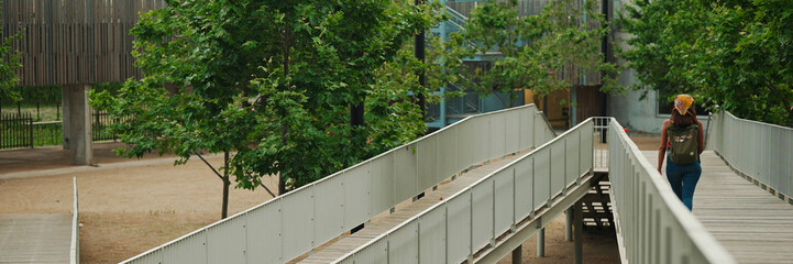 Fototapeta premium Back view, Cute tanned woman with long brown hair in white top and yellow bandana with backpack on her shoulders walks along the bridge, Panorama