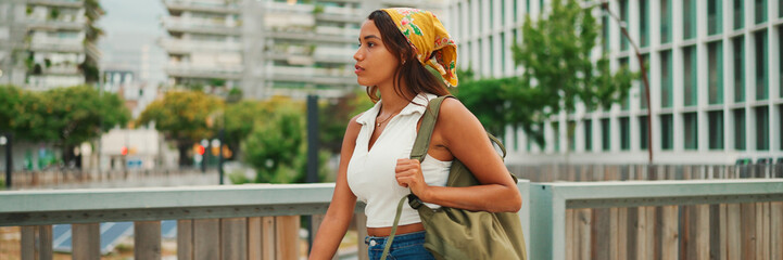 Сute tanned woman with long brown hair wearing white top and yellow bandana walks on bridge with...