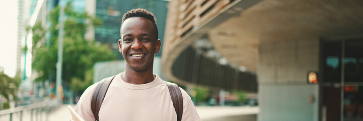 Young african student stands outside of university, looks at the camera and smiles, Panorama