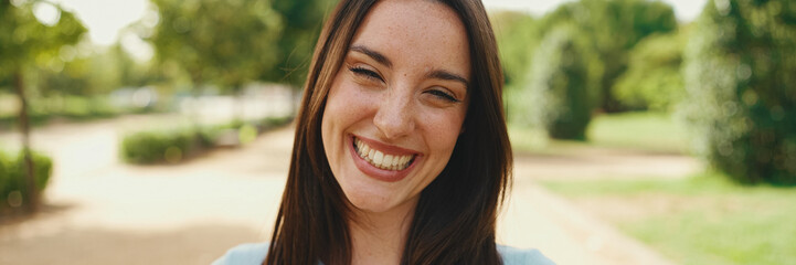 Close-up of young smiling woman with long brown hair wearing white crop top posing for the camera in the park, Panorama. The girl opens her eyes and smiles at the camera - obrazy, fototapety, plakaty