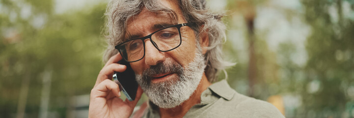 Panorama, Close up portrait od friendly middle aged man with gray hair and beard wearing casual clothes using his mobile phone. Mature gentleman in eyeglasses talking on cell phone outdoors - obrazy, fototapety, plakaty