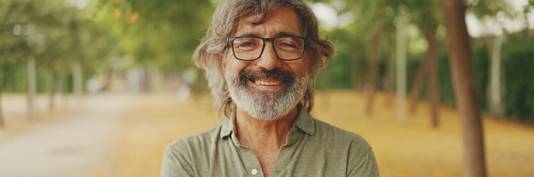 Fototapeta Clouse-up, friendly middle-aged man with gray hair and beard wearing casual clothes looks at the camera, Panorama. Mature gentleman in eyeglasses smiles at the camera