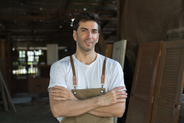 Portrait of male carpenter standing with crossed arms and smiling at woodwork workshop. Male joiner in furniture workshop. Start up and small business concept