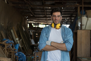 Portrait of male carpenter standing with crossed arms and smiling at woodwork workshop. Male joiner in furniture workshop. Start up and small business concept