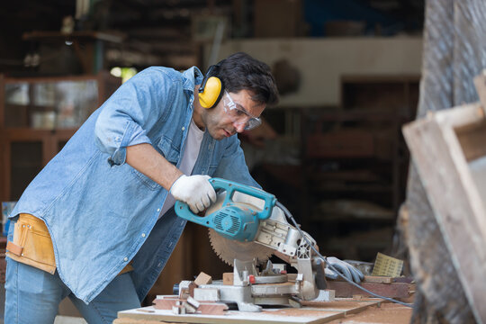 Male carpenter using electric saw cutting piece of wood and wearing safety goggles at woodwork workshop. Male joiner wearing safety equipment and working in furniture workshop