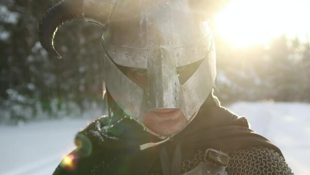 Medieval fantasy warrior in a horned helmet, posing against the backdrop of sunset in a winter forest. Rays of the sun, glare. Camera movement, selective focus.