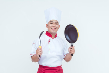 A cheerful Asian chef holding a frying pan and spatula spoon, dressed in traditional white chef attire, isolated on a white background.