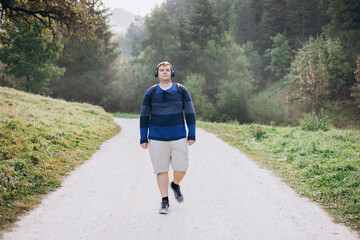 Active Young male with backpack and headphones hiking on the road in nature. 30s men walking on...