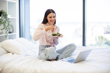 Beautiful young woman relaxing on cozy bed at home and using modern laptop while eating healthy salad. Caucasian brunette enjoying breakfast of fresh vegetables on weekend.