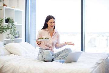 Happy brunette female talking by video call using laptop and eating healthy salad. Charming woman...