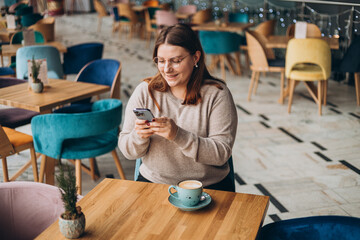 Young happy woman in glasses smiling and using smart phone in a cafe. Person sitting at table and using smartphone indoors. Online education, order, working or shopping concept