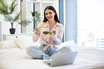 Portrait of young woman relaxing on cozy bad at home and using modern laptop while eating healthy salad. Caucasian brunette enjoying breakfast of fresh vegetables on weekend, looking at camera.
