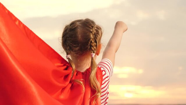 Cute baby girl superhero costume red cloak flying at sunset sunrise sky back view closeup. Funny adorable female kid child in superman costume forward movement at cloud imagination and freedom hero