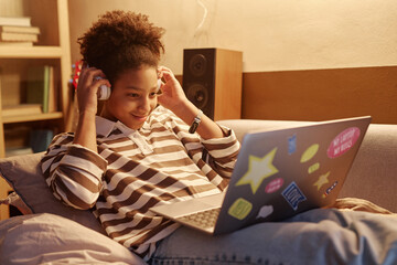 Portrait of smiling Black teenage girl using laptop with wireless headphones at home at night