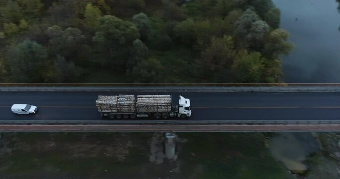 Double length log semi truck driving on bridge on straight highway. Freight truck haul timber production across asphalt road. Aerial drone top down follow mid shot at summer sunset