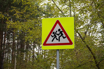Road sign Denoting Be careful, children, on pole, that is installed near schools. Yellow square, red triangle, white, with the image of running people. Safety, Traffic warning, autumn forest.