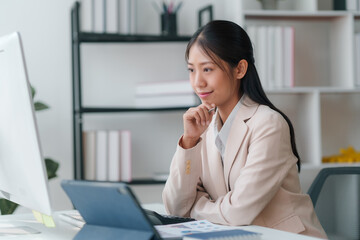 Professional young asian businesswoman smiling while focusing on data analysis on her desktop computer in a modern office.