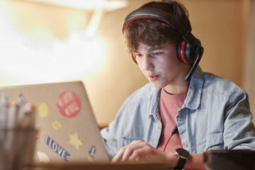 Portrait of teenage boy playing video games online on laptop with wireless headset at night in...