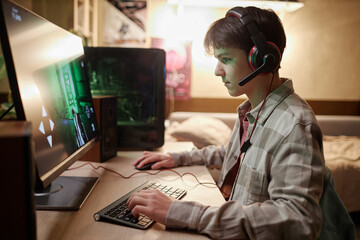 Side view portrait of teenage boy playing video games online on PC computer with wireless headset...
