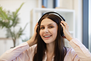Portrait of happy pretty woman using modern device listening favorite music at home. Happy lady in headphones sitting on comfy bed and enjoying relaxing music on weekend.