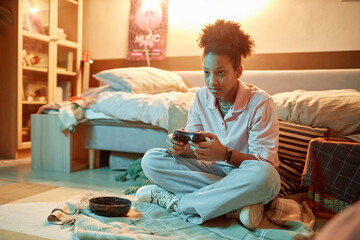 Portrait of African American teen girl holding gamepad and playing videogames sitting on floor at...