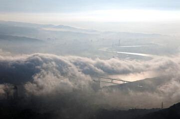 Naklejka premium Aerial panorama of Taipei City on a foggy morning with a bird's eye view of dense fog rolling over Guandu Plain, a bridge across Tamsui River & mountain silhouettes in the background