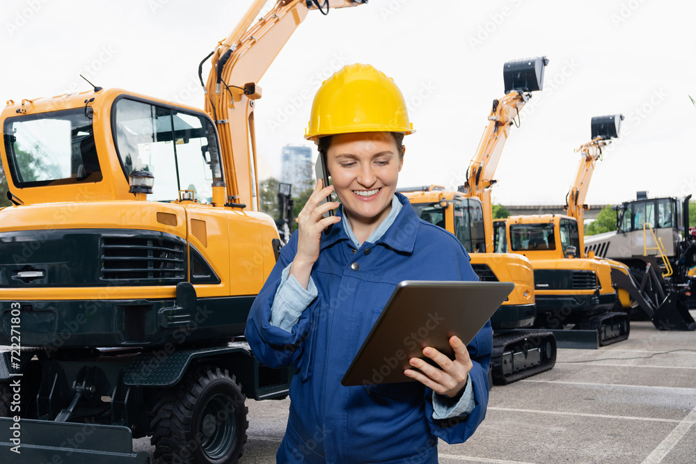 Poster engineer in a helmet with a digital tablet stands next to construction excavators