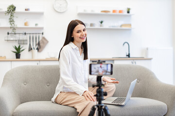 Front view of cheerful content maker in casual wear recording video to followers on modern smartphone. Young caucasian woman in with laptop sitting on comfortable sofa in kitchen.