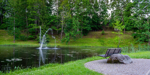 Bench to relax in a shady green park with fountain in the pond. Smiltene Old Park, Latvia.