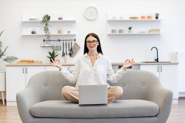 Caucasian woman meditating with closed eyes while working on portable laptop at living room. Brunette female freelancer sitting on comfy couch in lotus pose with reducing fatigue using yoga practice.