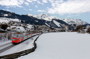 A local train traveling by Salzach River through the snowy valley on a sunny winter day, with...