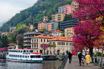 Tourists walk on a lakeside promenade under beautiful blossoming trees by Lake Como in Lombardy Italy, with ferry boats parking at the dock & colorful houses standing along lakeshore & on the hillside