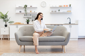 Obraz premium Concept of unification people and technology. Positive brunette young female in casual attire gesturing with hands while sitting on couch with modern laptop and looking at web camera.
