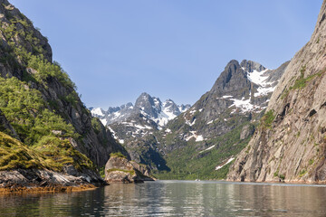 A bright summer day in Lofoten, Norway, where an excursion catamaran glides on Trollfjorden's reflective water, flanked by steep, verdant cliffs and snow-patched mountains