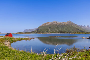 A serene Norwegian landscape showcases a traditional red boathouse on the water's edge with a majestic mountain backdrop under the clear blue skies of Lofoten