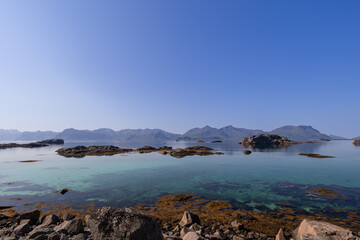 Fototapeta na wymiar Crystal-clear waters meet the rocky shores of Lofoten, offering a tranquil view of distant mountains under the vast expanse of the Nordic sky