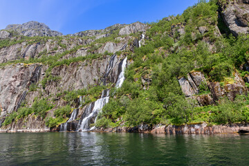 A bright summer day in Lofoten unveils a spectacular waterfall spilling over the cliffs into the clear waters of Trollfjorden, Norway.